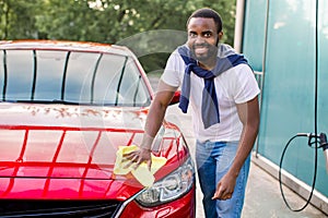 Young happy smiling African man cleaning red car hood and headlight with yellow microfiber cloth, looking at camera