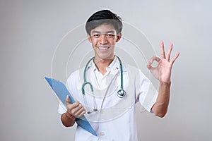 Young happy smiling Asian medical doctor holding medical chart with okay gesture over gray studio background