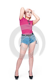 Young happy smiling girl in denim shorts and a pink t-shirt stands in high heels. Isolated on white background.