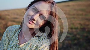 Young happy smiling lady with long hair in floral dress being in the field turning towards the sun and closes it with