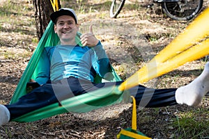 Young happy smiling man in hat relaxing outside in hammock in forest.