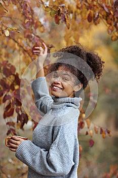 Young happy smiling mixed-race woman with coffee cup in autumn nature, pleased african american female with curly hair in knitted