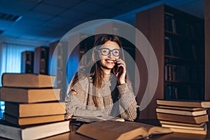 Young happy smiling student in glasses preparing for the exam. Girl in the evening sits at a table in the library with a pile of