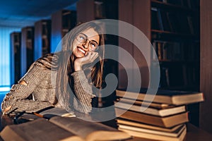 Young happy smiling student in glasses preparing for the exam. Girl in the evening sits at a table in the library with a pile of