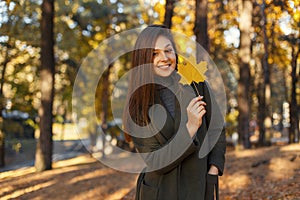 Young happy smiling stylish girl in a fashionable green coat holding a golden autumn leaf walks in the park on a sunny day.