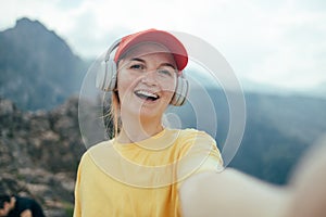 Young happy smiling woman backpacker with short brunette hair and yellow beanie, taking selfie with smartphone doing