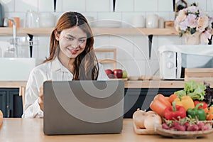 Young happy smiling woman using laptop while cooking vegan food in the modern kitchen, smiling hipster girl preparing