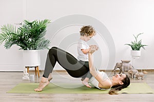 Young happy sporty woman mother working out, doing butt bridge pilates yoga exercise, little kid son on her belly