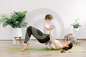 Young happy sporty woman mother working out, doing butt bridge pilates yoga exercise, little kid son on her belly photo