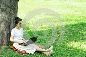 Young happy student asian woman sitting on grass at park working on laptop,relaxing with coffee while under a tree.Nature