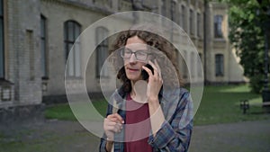 Young happy student with curly hair going to college and talking on phone, walking in park near university, daytime