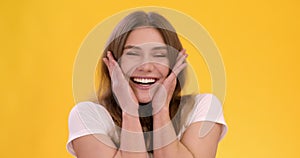 Young happy surprised woman shouting in amazement and delight, touching her cheeks, yellow studio background