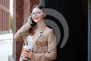 Young happy teen girl in beige stylish coat speaking on cellphone while standing on city street