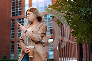 Young happy teen girl in beige stylish coat speaking on cellphone while standing on city street