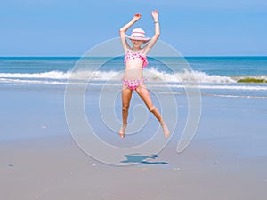 Young happy teen girl having fun on tropical beach and jumping in pink swimsuit and striped hat into the air on the sea coast