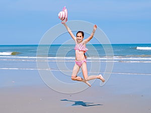 Young happy teen girl having fun on tropical beach and jumping in pink swimsuit and striped hat into the air on the sea coast