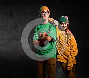 Young happy teens boy and girl in green and yellow stylish casual clothing and hats standing and smiling over dark background