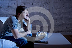 Young happy television addict man sitting on home sofa watching TV and eating popcorn