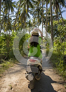 Young happy tourist woman with hat riding scooter motorbike in tropical paradise jungle with blue sky and palm trees exploring tr