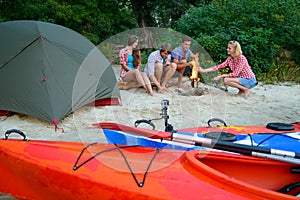 Young Happy Travelers Resting at Evening with Fire on the Sand Beach near Kayaks and Tent.