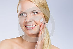 Young happy woman applying cream on face looking away