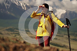 Young happy woman with backpack standing on a rock and looking through binocular to a valley below.