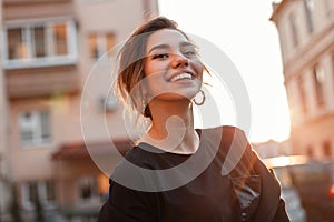 Young happy woman with a beautiful smile in a stylish T-shirt in a coat is standing and enjoying the orange sunset in the city