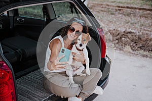 Young happy woman in a car enjoying with her cute dog. Taking a selfie with mobile phone. Travel concept