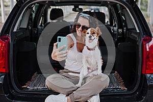 Young happy woman in a car enjoying with her cute dog. Taking a selfie with mobile phone. Travel concept