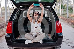 Young happy woman in a car enjoying with her cute dog. Taking a selfie with mobile phone. Travel concept