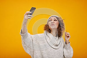 Young happy woman with curly hair in winter hat standing over yellow background making selfie. Happy New Year and Christmas