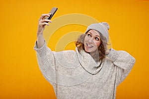 Young happy woman with curly hair in winter hat standing over yellow background making selfie. Happy New Year and Christmas