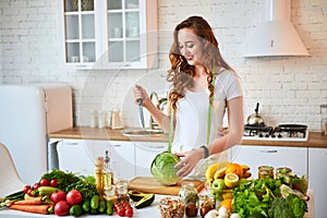Young happy woman cutting cabbage for making salad in the beautiful kitchen with green fresh ingredients indoors. Healthy food and