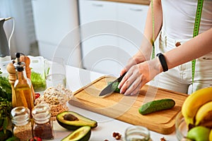 Young happy woman cutting cucumber for making salad in the beautiful kitchen with green fresh ingredients indoors. Healthy food