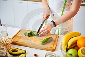 Young happy woman cutting cucumber for making salad in the beautiful kitchen with green fresh ingredients indoors. Healthy food