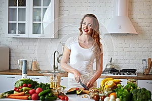 Young happy woman cutting tomatoes for making salad in the beautiful kitchen with green fresh ingredients indoors. Healthy food