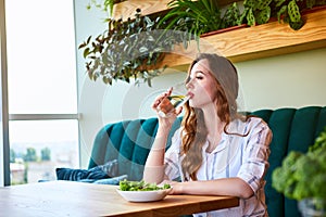 Young happy woman drinks water in the beautiful interior with green flowers on the background and fresh ingredients on the table.