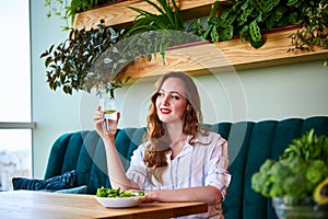 Young happy woman drinks water in the beautiful interior with green flowers on the background and fresh ingredients on the table.