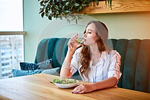 Young happy woman drinks water in the beautiful interior with green flowers on the background and fresh ingredients on the table.