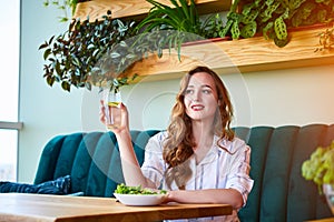 Young happy woman drinks water in the beautiful interior with green flowers on the background and fresh ingredients on the table.