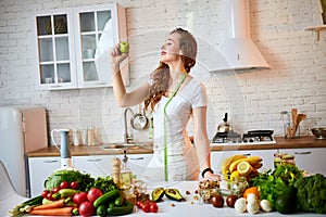 Young happy woman eating apple in the beautiful kitchen with green fresh ingredients indoors. Healthy food and Dieting concept.