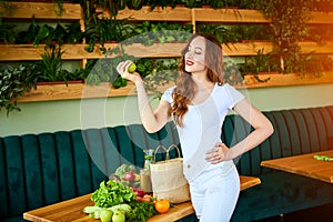 Young happy woman eating apple in the beautiful kitchen with green fresh ingredients indoors. Healthy food and Dieting concept.