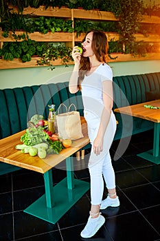 Young happy woman eating apple in the beautiful kitchen with green fresh ingredients indoors. Healthy food and Dieting concept.