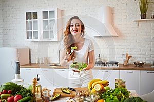 Young happy woman eating salad in the beautiful kitchen with green fresh ingredients indoors. Healthy food concept