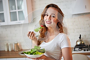 Young happy woman eating salad in the beautiful kitchen with green fresh ingredients indoors. Healthy food concept