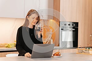 Young happy woman enjoys her morning coffee while checking laptop in kitchen.