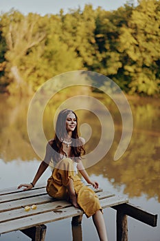 A young happy woman with a hippie smile sits on the lake shore on a bridge wearing eco clothing made of natural