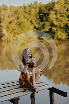 A young happy woman with a hippie smile sits on the lake shore on a bridge wearing eco clothing made of natural