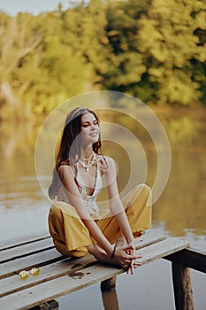 A young happy woman with a hippie smile sits on the lake shore on a bridge wearing eco clothing made of natural