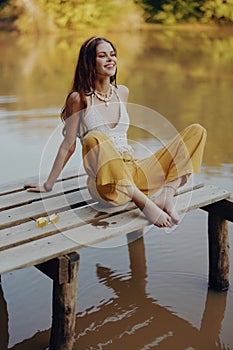 A young happy woman with a hippie smile sits on the lake shore on a bridge wearing eco clothing made of natural